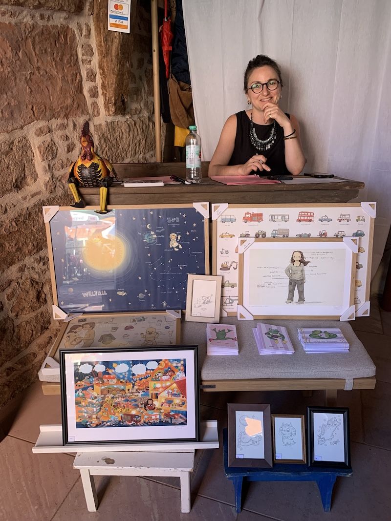 Marie-Cécile am Tresen des Pop-up-Stores, davor gerahmte Illustrationen und Postkarten